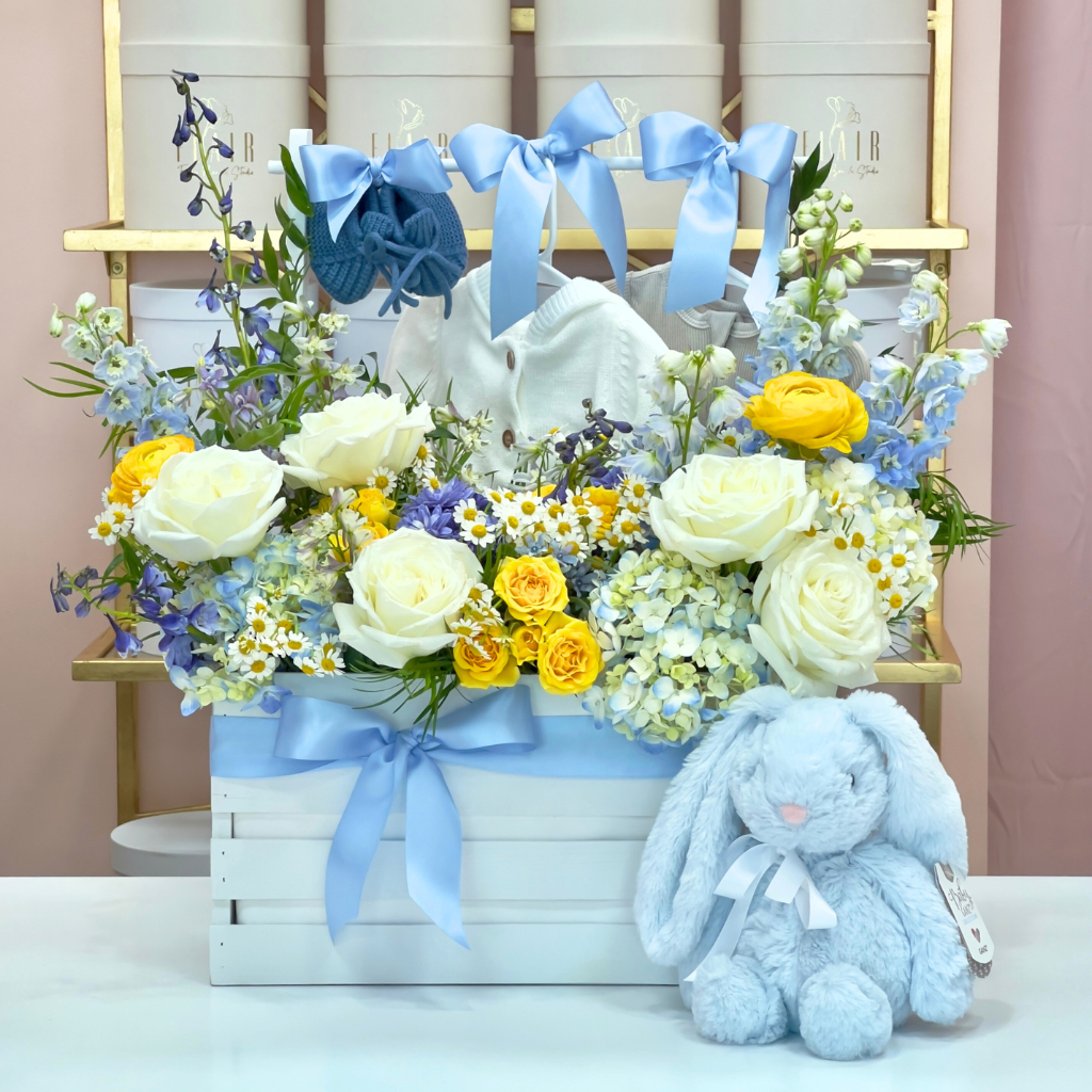 Floral arrangement in a basket with blue ribbons and a plush bunny toy on a white surface.