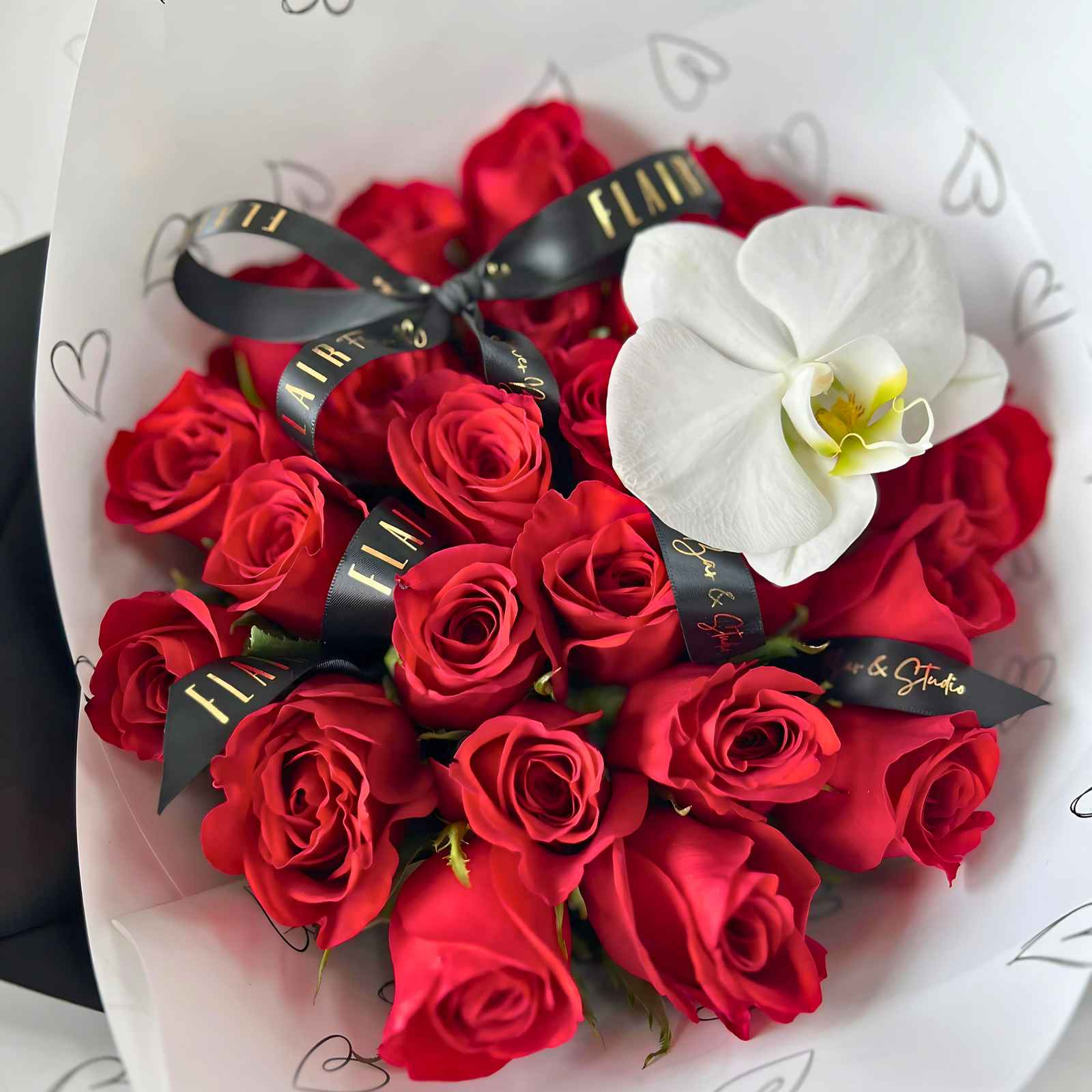 A round, dome-like bouquet of red roses with a white orchid on top, elegantly wrapped with a black ribbon and a signature sticker.