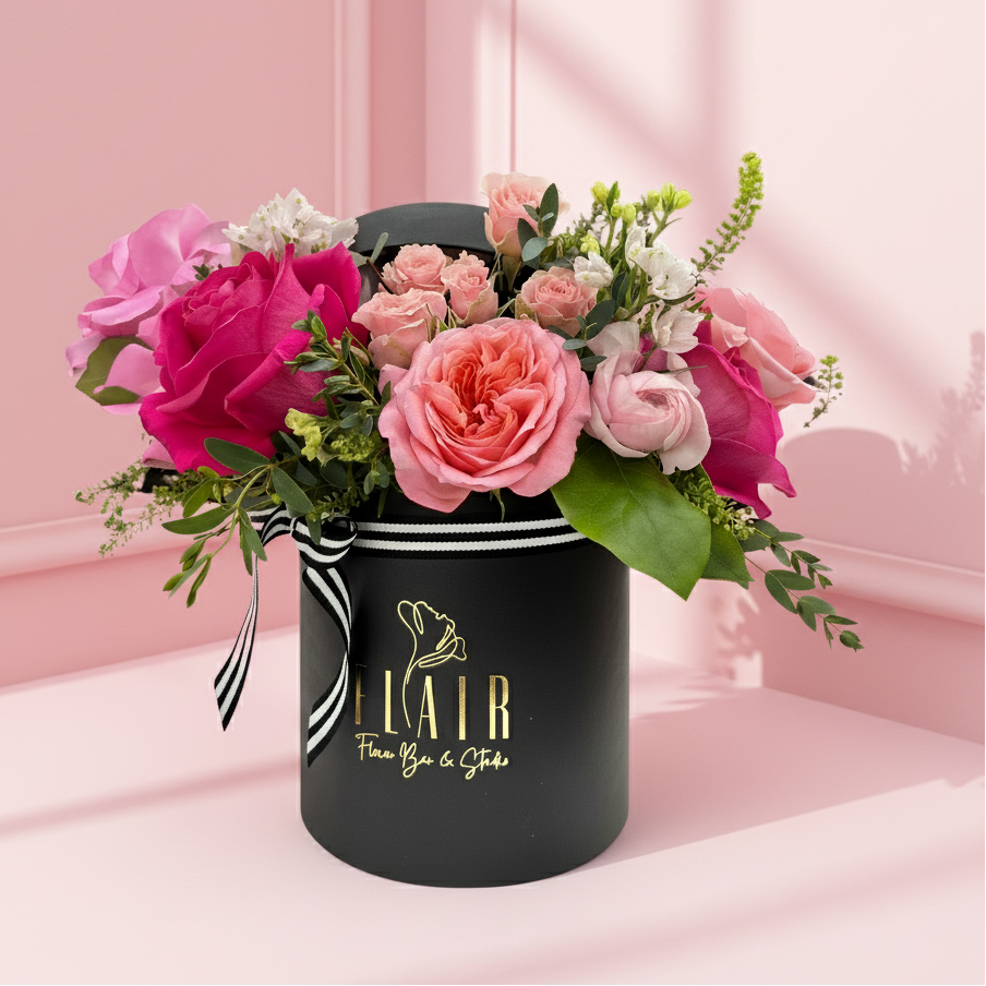 A floral arrangement with pink and red roses, green foliage, and a black-and-white striped ribbon in a round black box with the logo 'FLAIR' on the front.