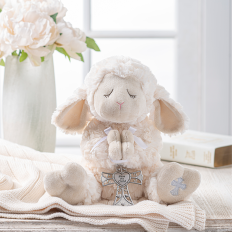 Plush lamb toy with a cross on a wooden surface with flowers and books in the background