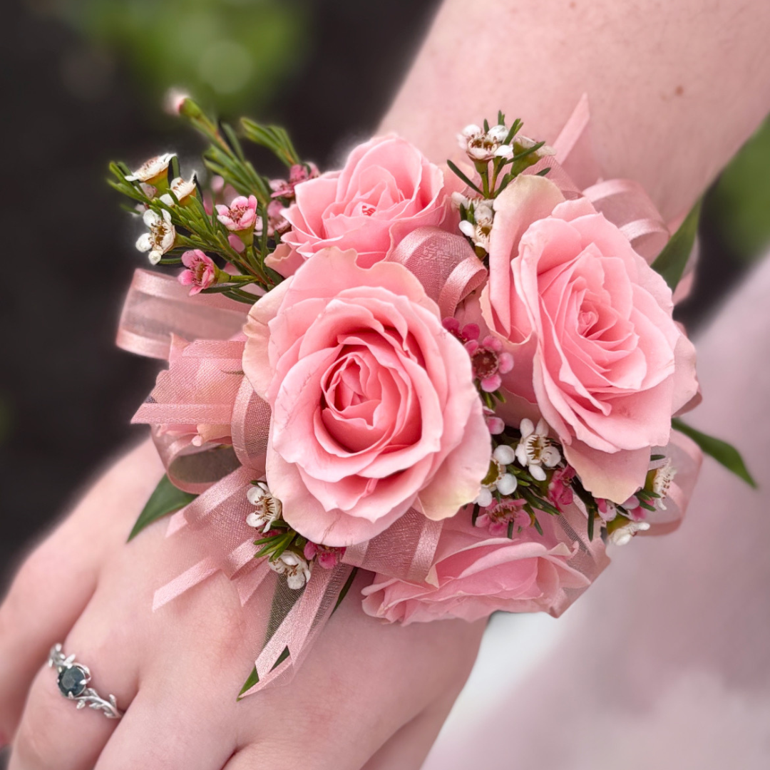 A wrist corsage featuring pink spray roses, white accent florals, and greenery, attached to a white rhinestone band.