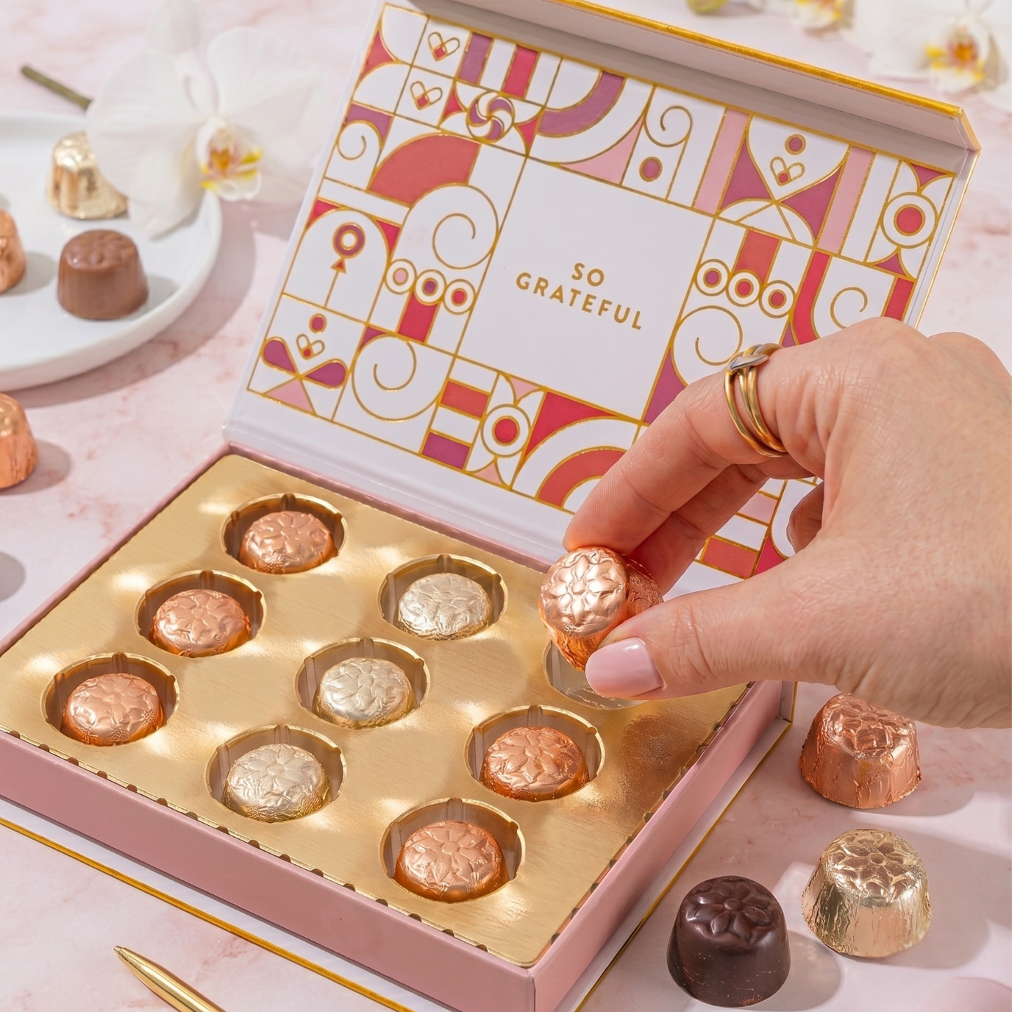 Hand reaching into a box of chocolates with a decorative cover on a light pink surface.