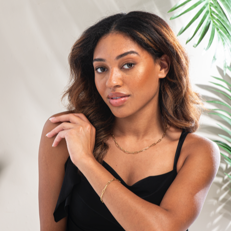 Woman in a black top with a plant in the background with gold bracelet and necklace