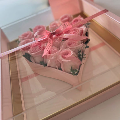 Heart-shaped box of pink roses with a pink ribbon on a reflective surface.