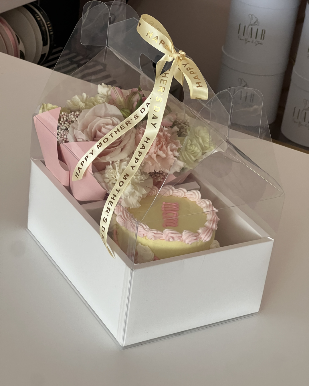 Gift set with flowers and cake in a clear gift box on a white surface