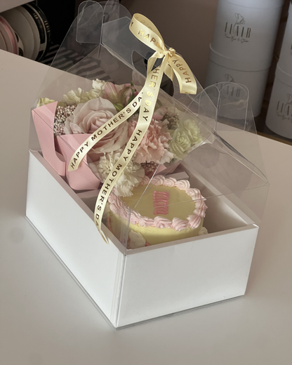 Gift set with flowers and cake in a clear gift box on a white surface