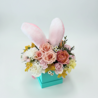 Floral arrangement with pink bunny ears in a turquoise box on a light gray background