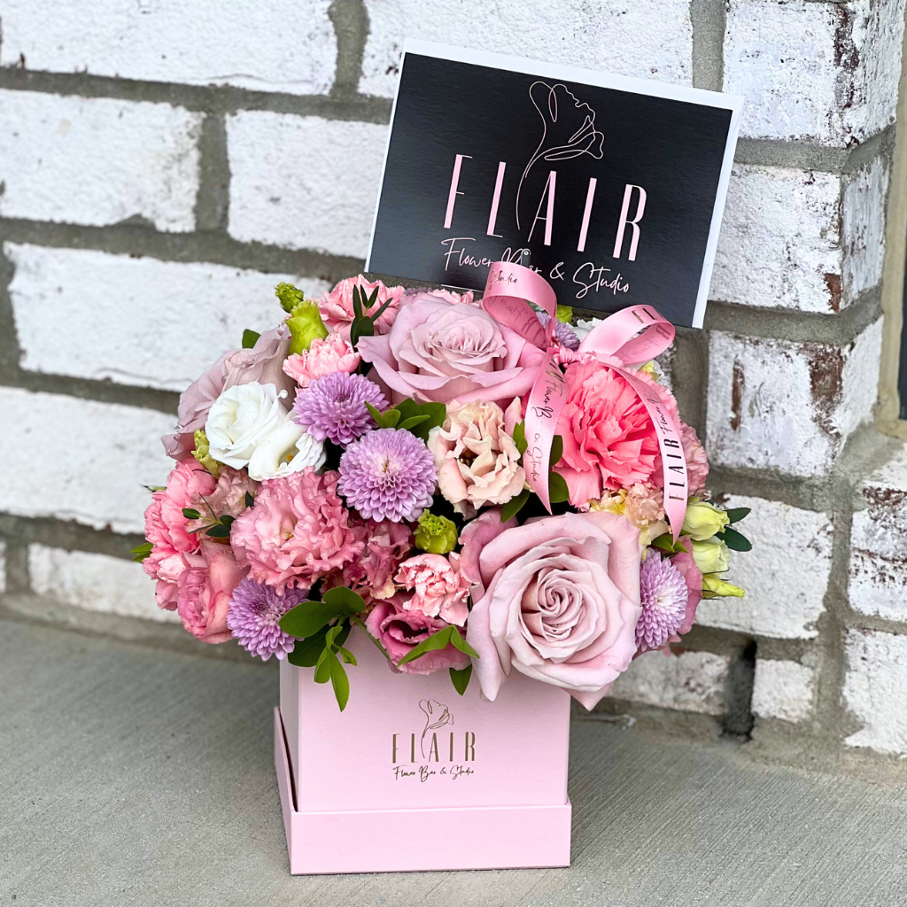 Floral arrangement in a pink box with 'FLAIR' branding against a brick wall.