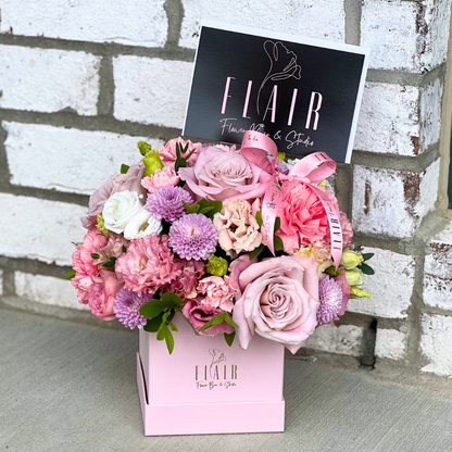 Floral arrangement in a pink box with 'FLAIR' branding against a brick wall.