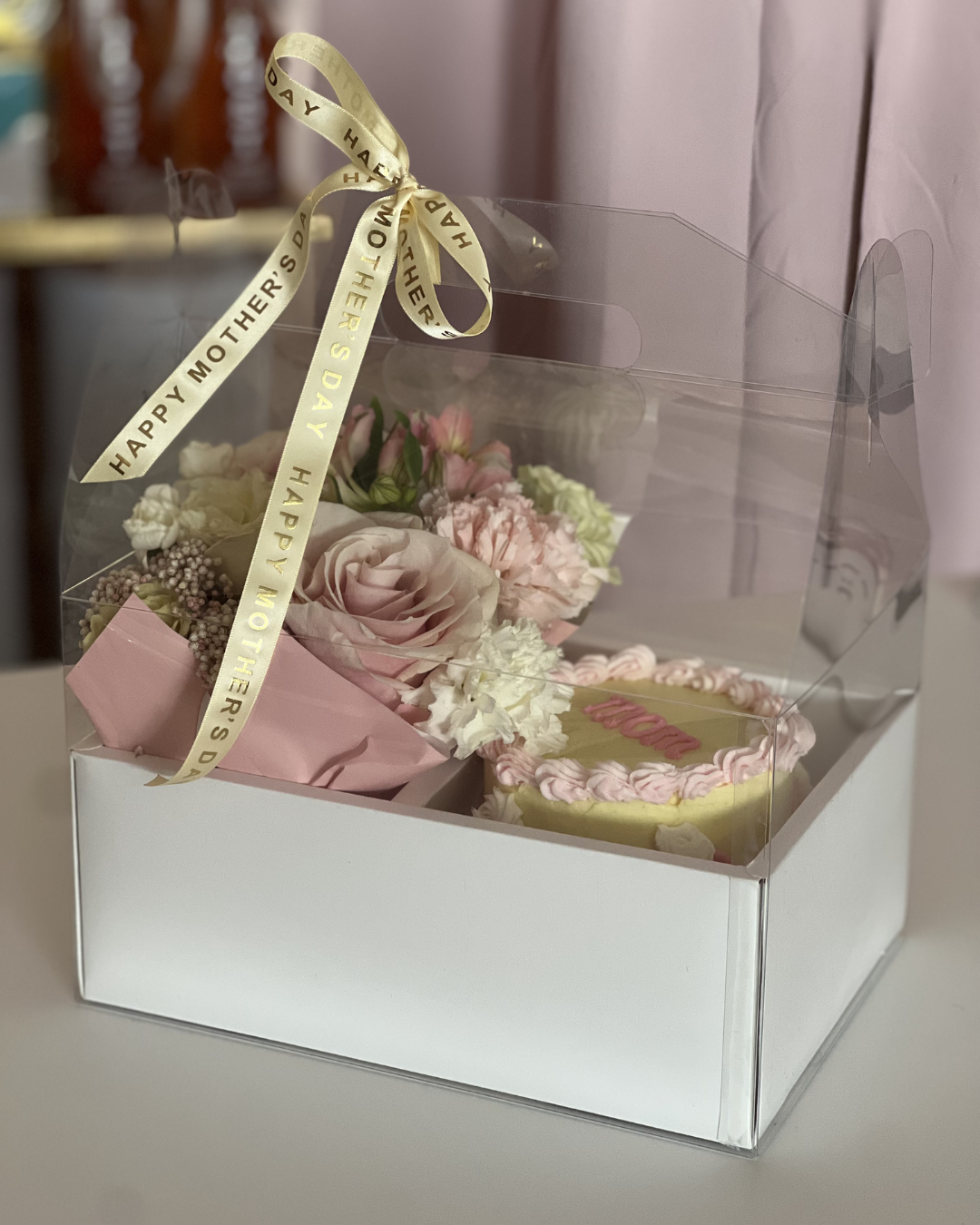 Gift box with flowers and a cake, tied with a 'Happy Mother's Day' ribbon. 