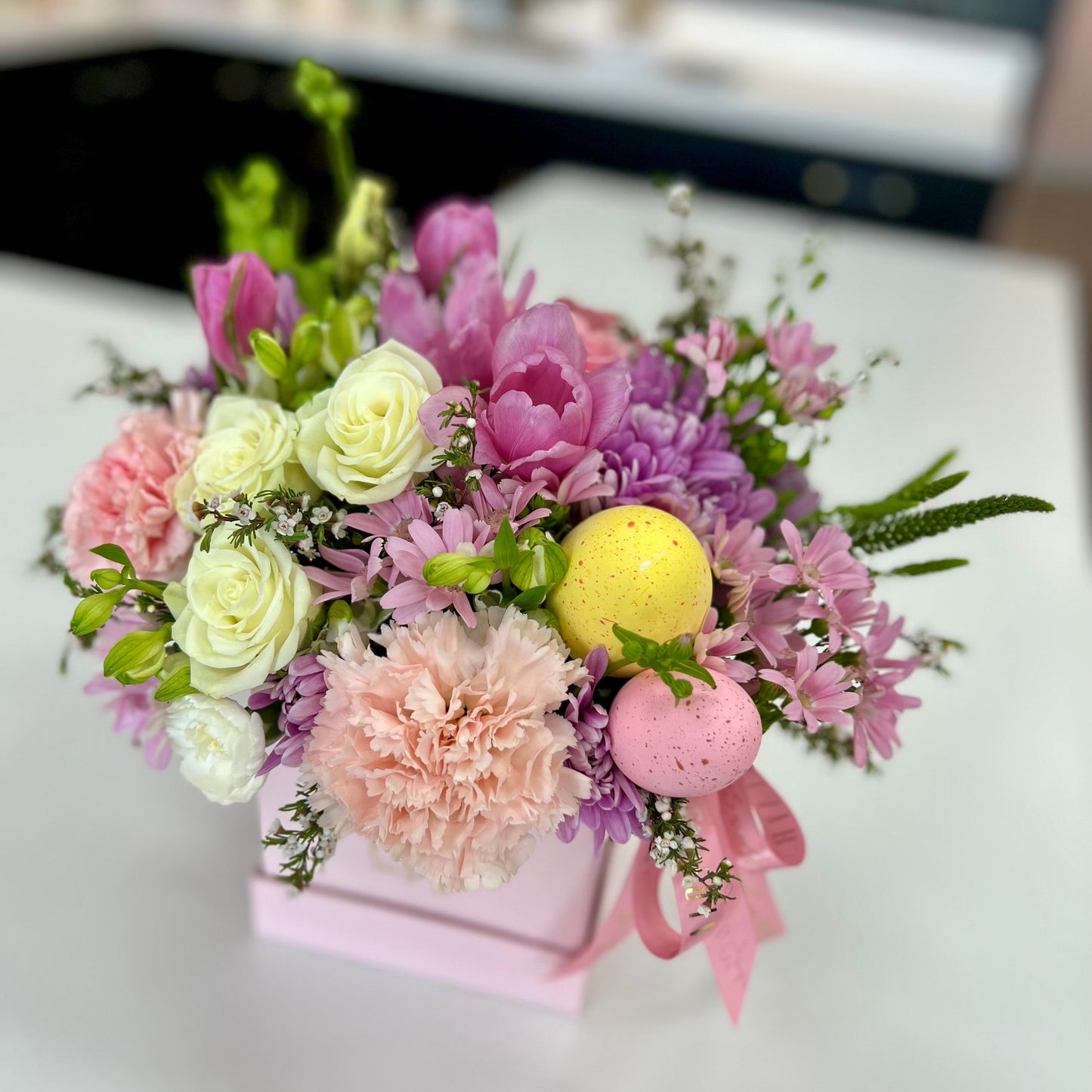 Floral arrangement with pastel-colored flowers and decorative eggs in a pink box on a white surface.