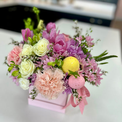Floral arrangement with pastel-colored flowers and decorative eggs in a pink box on a white surface.