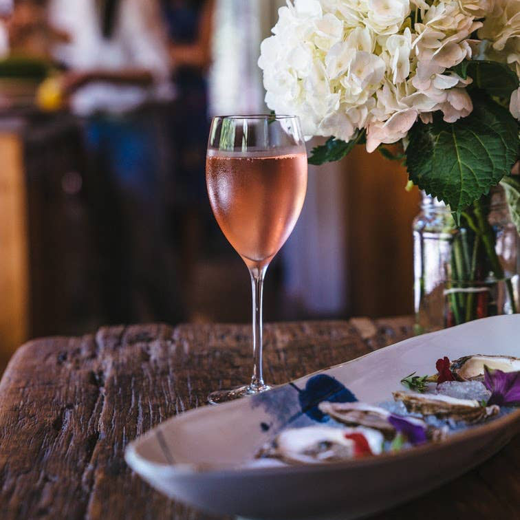 Glass of pink wine on a table with a plate of food and a bouquet of flowers in the background.