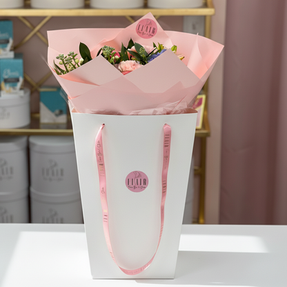 The Maison Wrap Floral arrangement in a white box with pink handles and pink paper, on a white surface with a blurred background.