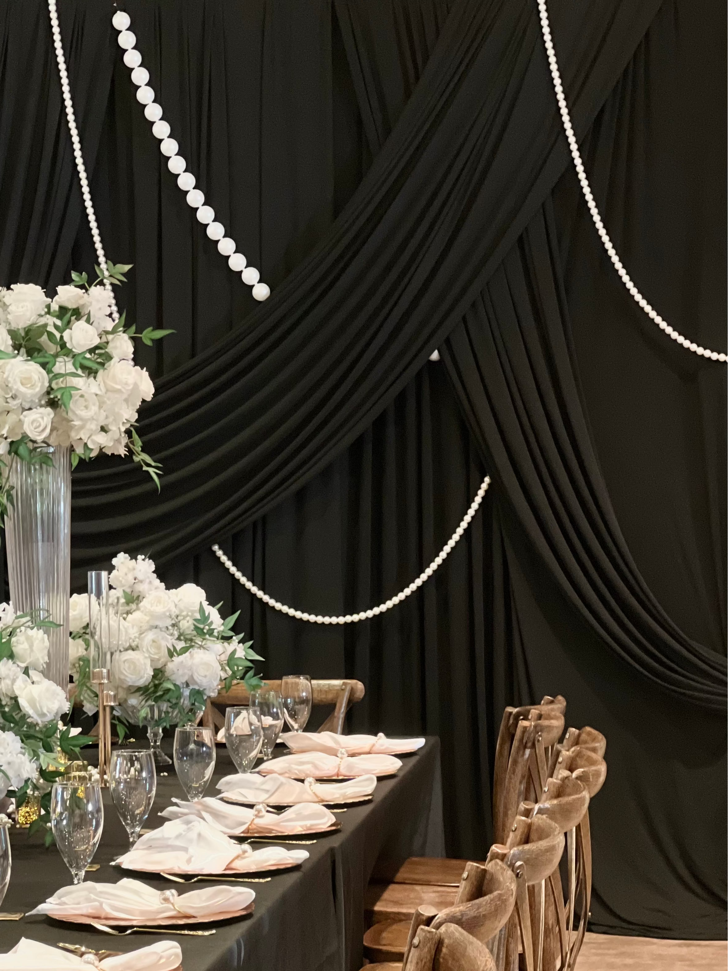 A long dining table set for an event with black tablecloths, folded white napkins, clear glassware, and large white floral arrangements. Wooden chairs line one side of the table, and the background features black draped fabric with hanging pearl bead garlands.