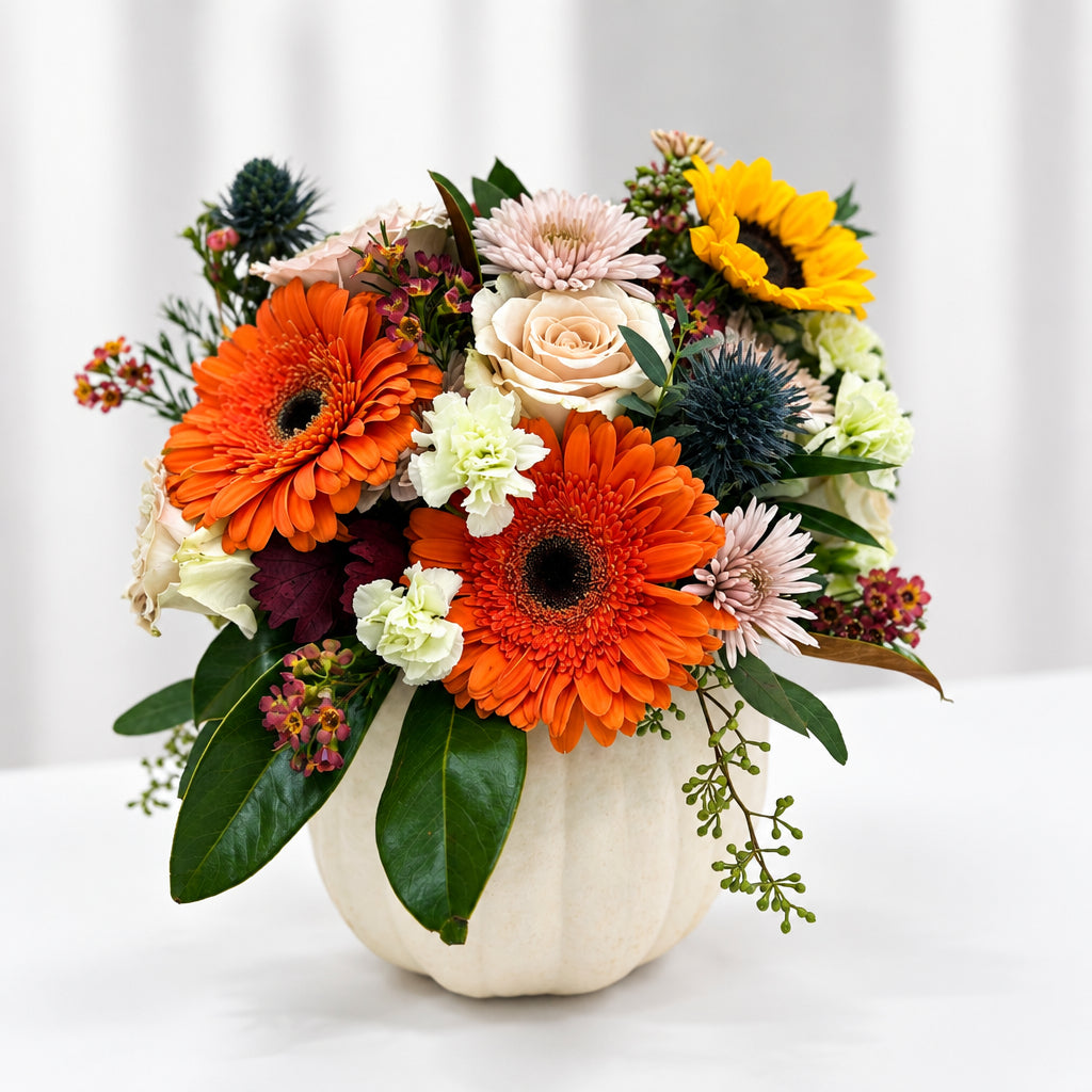 A vibrant autumn floral arrangement featuring orange gerbera daisies, yellow sunflowers, cream roses, and green foliage, presented in a small white real pumpkin on a white table.