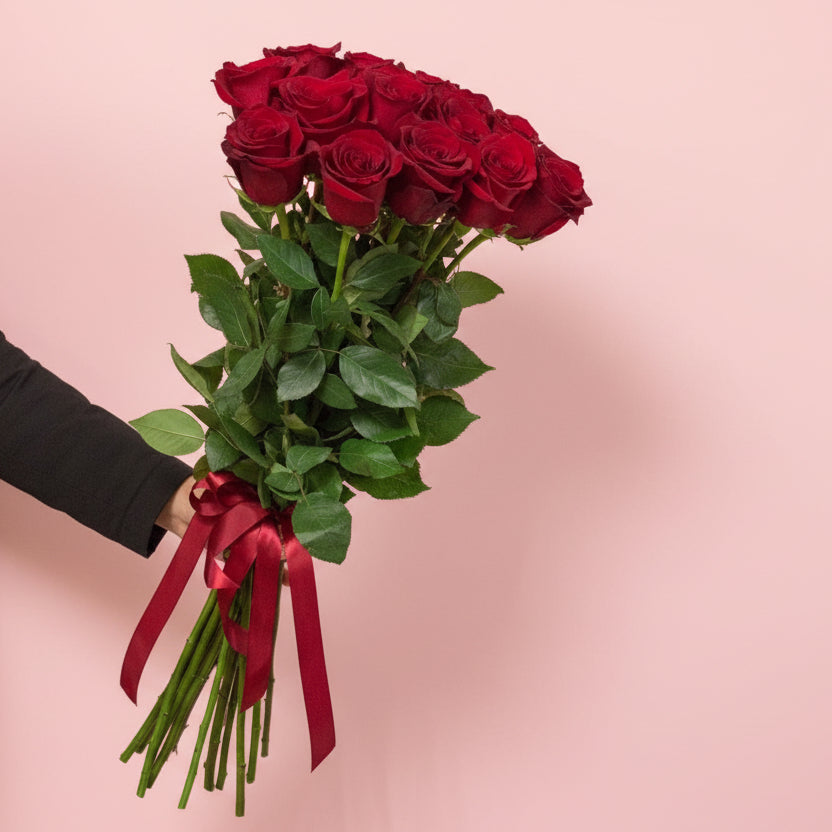 Bouquet of red roses with a red ribbon held by a person against a pink background - Flair Flower Bar