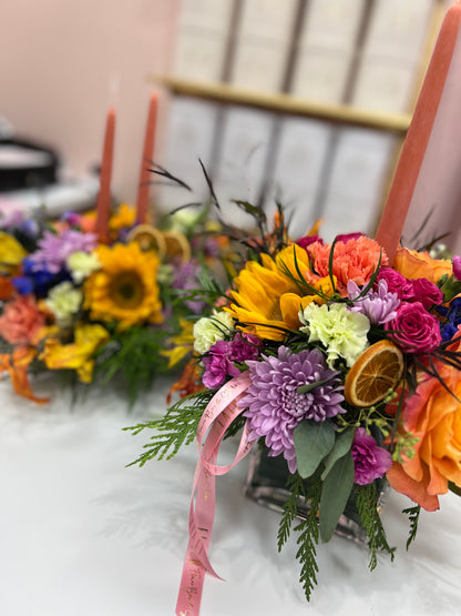 A close-up of a colorful floral centerpiece arrangement in a clear glass vase, featuring orange roses, purple chrysanthemums, yellow sunflowers, green foliage, dried orange slices, pink candles, and a pink ribbon, set on a white table with a softly blurred indoor background.