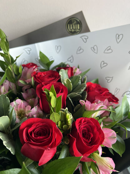 A close-up of a flower bouquet featuring red roses and pink alstroemeria flowers with green foliage, wrapped in white paper with small heart outlines and available for local Cincinnati delivery by Flair Flower Bar in Florence, Kentucky