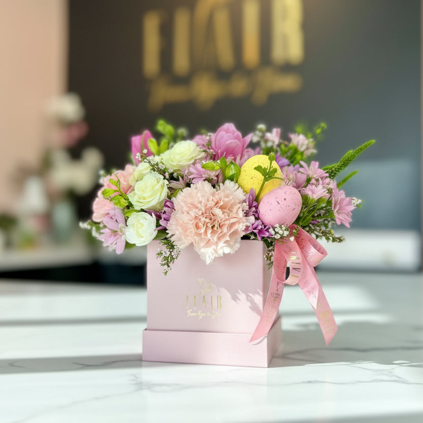 Floral arrangement in a pink box with a pink ribbon, featuring a blurred background with a logo.