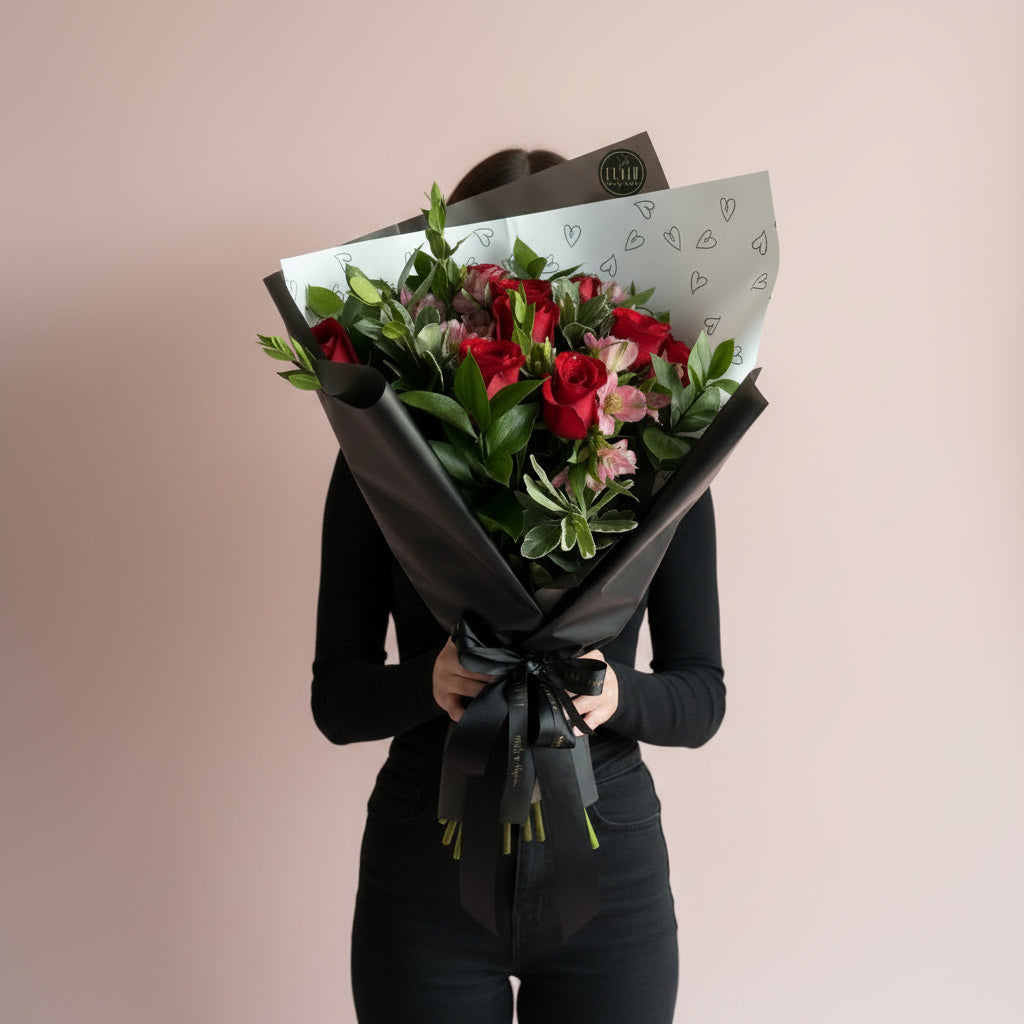 A woman holding a fresh flower bouquet wrapped in black paper, featuring 12 red roses, pink flowers, and green leaves, with a white patterned insert paper. The bouquet is held in front of a plain beige wall next to a window.