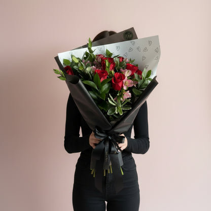 A woman holding a fresh flower bouquet wrapped in black paper, featuring 12 red roses, pink flowers, and green leaves, with a white patterned insert paper. The bouquet is held in front of a plain beige wall next to a window.
