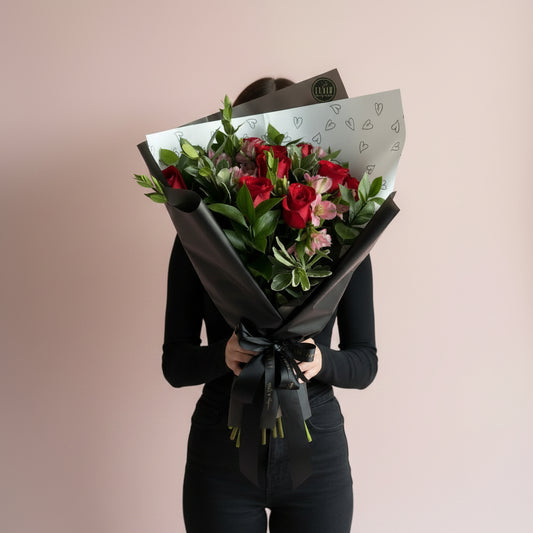 A woman holding a fresh flower bouquet wrapped in black paper, featuring 12 red roses, pink flowers, and green leaves, with a white patterned insert paper. The bouquet is held in front of a plain beige wall next to a window.