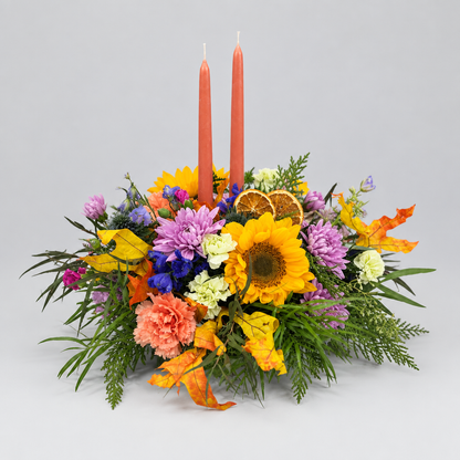 A vibrant floral centerpiece arrangement featuring sunflowers, carnations, purple flowers, green foliage, dried orange slices, and two tall orange taper candles, photographed on a neutral light gray background for Cincinnati and Northern Kentucky from Flair Flower Bar.