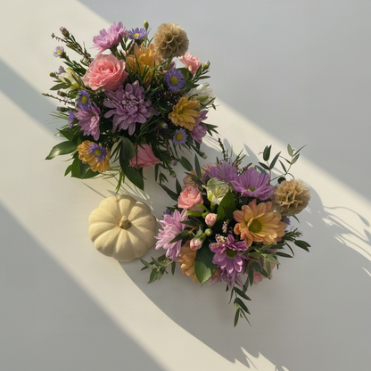 Two pumpkin fall floral arrangements with purple, pink, yellow, and green flowers on a white surface, styled with a small cream-colored pumpkin.