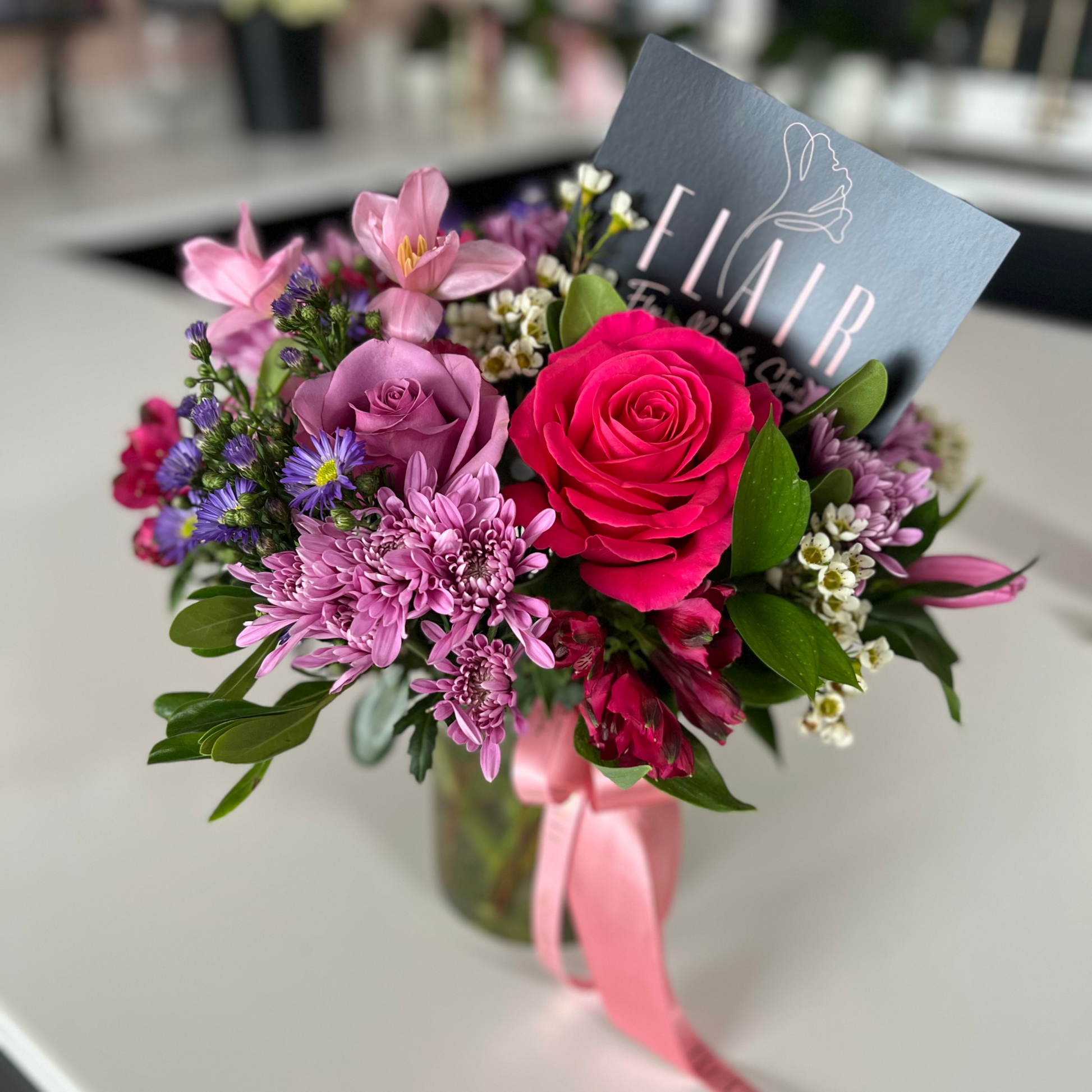 Bouquet of pink and purple flowers with a pink ribbon on a white surface, featuring the brand 'Flair'.
