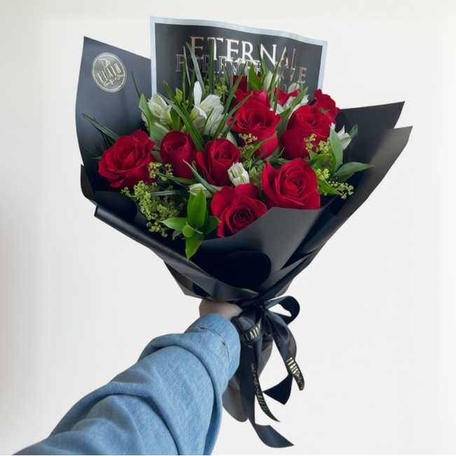 A hand wrapped bouquet of red roses and white lilies wrapped in black paper, with a card reading 'ETERNAL FOREVER LOVE' in the background by Flair Flower Bar in Florence, Ky