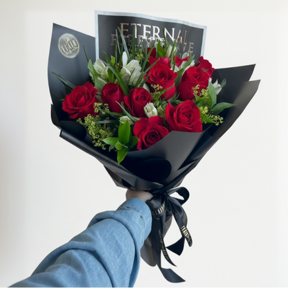 A hand wrapped bouquet of red roses and white lilies wrapped in black paper, with a card reading 'ETERNAL FOREVER LOVE' in the background by Flair Flower Bar in Florence, Ky