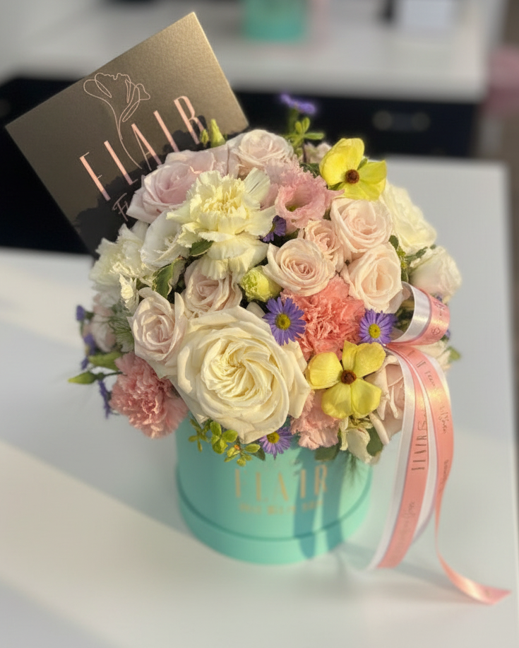 A floral arrangement in a round mint box with the brand name FLAIR visible on the box and card. The bouquet includes cream, blush pink, peach, yellow, and purple flowers, with a pink ribbon tied around the arrangement in Cincinnati and Florence, Kentucky