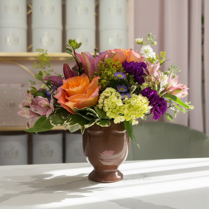 A vibrant floral arrangement in a brown ceramic vase, featuring orange roses, purple flowers, yellow hydrangeas, and pink blossoms, set on a white table with a pink curtain and shelves in the background by Flair Flower Bar & Studio in Florence, Kentucky