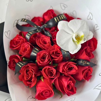 A round, dome-like bouquet of red roses with a white orchid on top, elegantly wrapped with a black ribbon and a signature sticker.