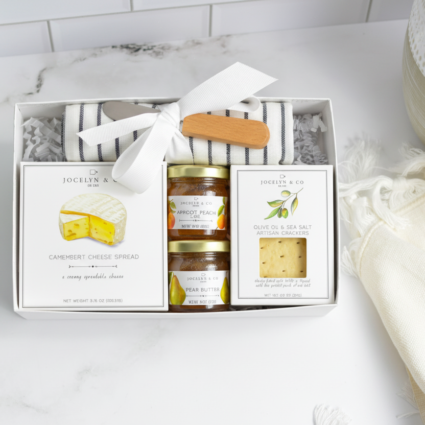 Charcuterie gift set with jars and a box on a marble surface with a plant and towel in the background.