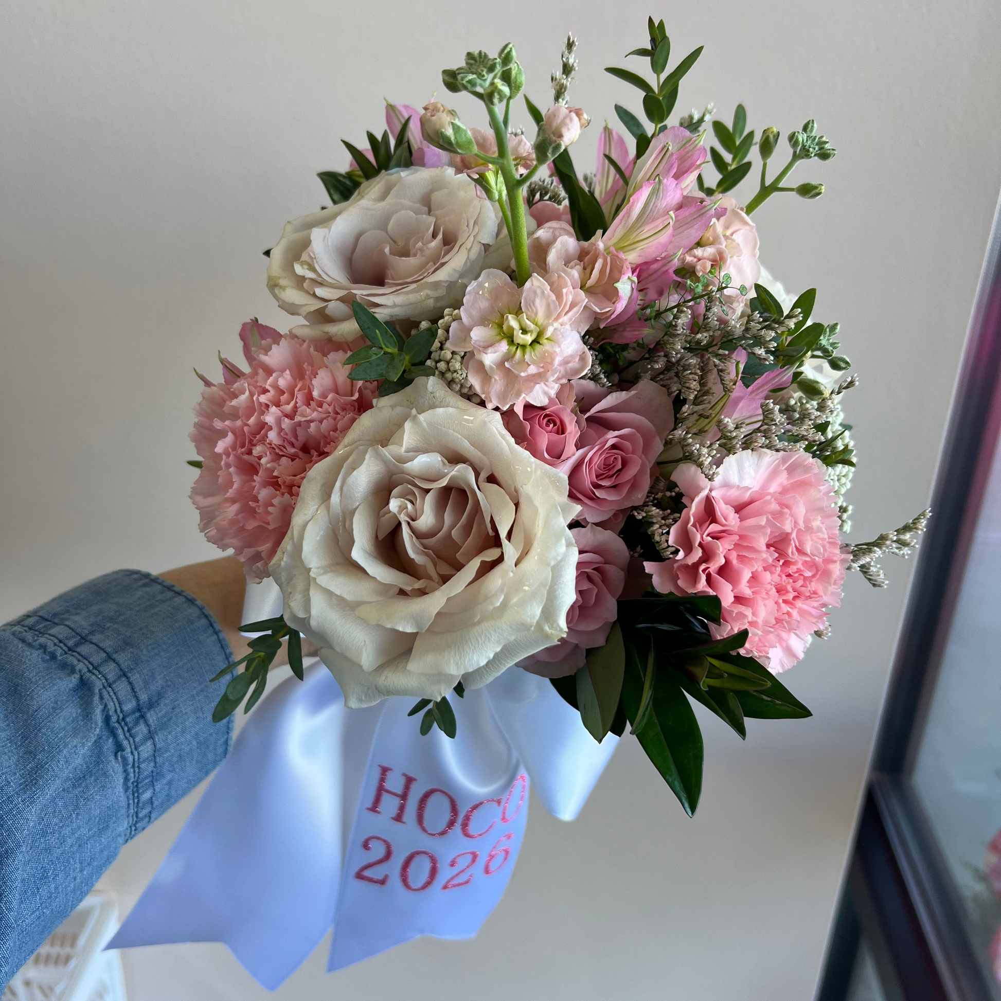 A hand holding a bouquet of pink and cream roses, carnations, and green foliage wrapped in a white bow ribbon with pink text reading 'HOCO 2026'.