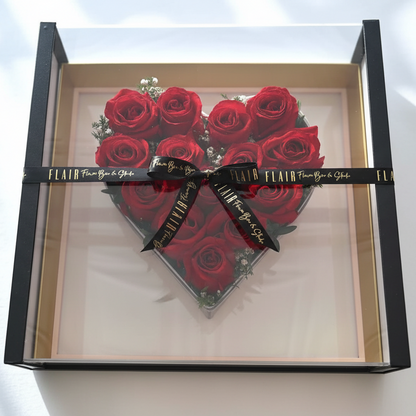 Heart-shaped arrangement of red roses in a clear box with a black ribbon on a white background