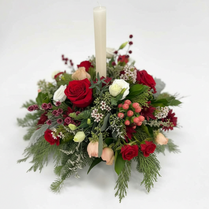 A Christmas candle centerpiece arrangement featuring a variety of flowers including red roses, white flowers, and greenery, topped with a single candle.