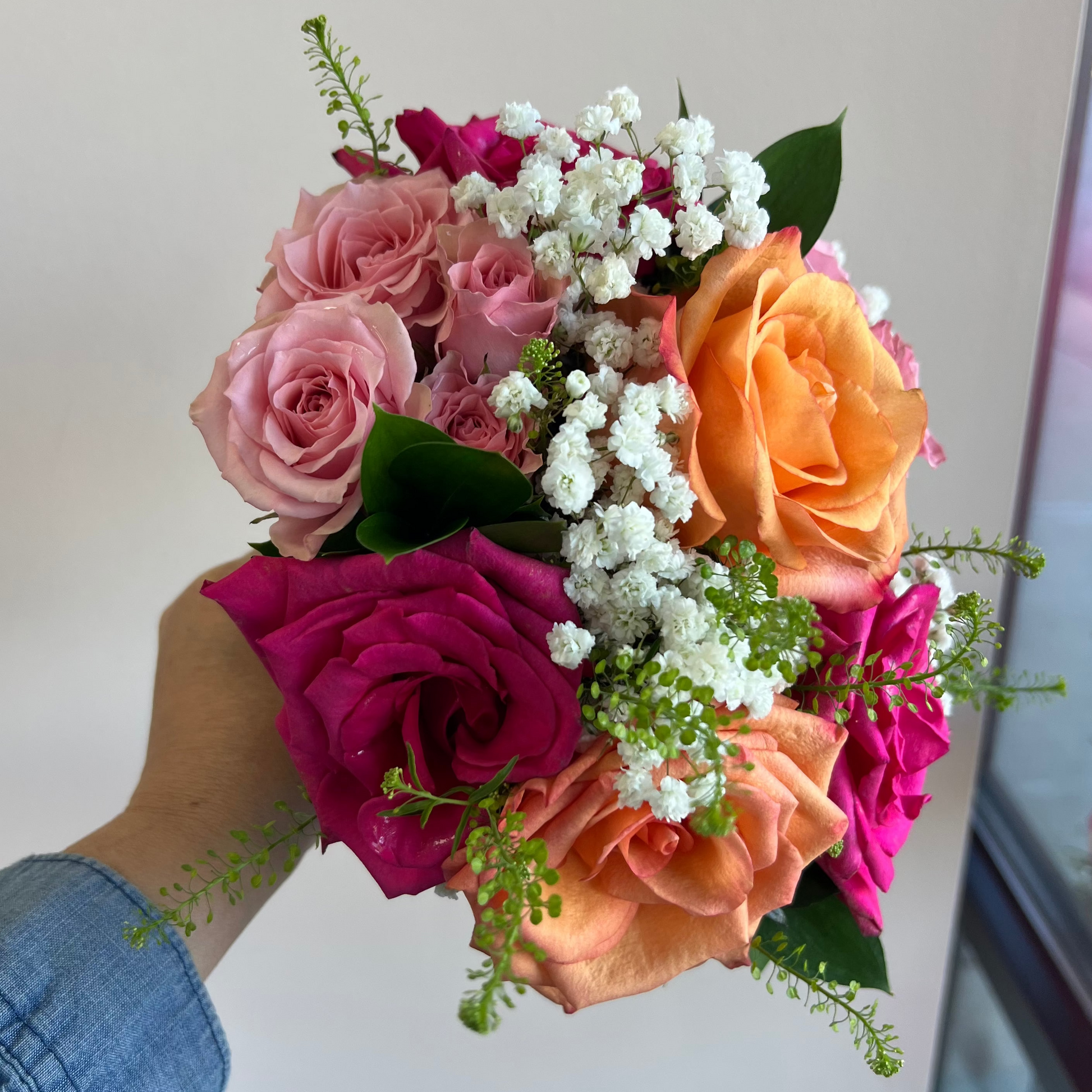 A hand holding a vibrant homecoming bouquet of roses in shades of pink, orange, and magenta, accented with white baby's breath and green foliage, set against a plain indoor wall.