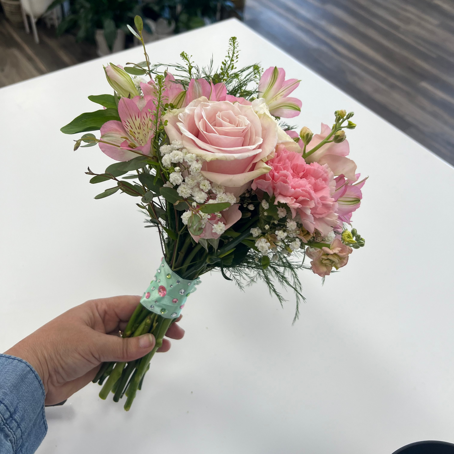 A hand holding a small bouquet of pink flowers, including a pink rose, pink carnations, and pink alstroemeria, with green foliage and white baby's breath, wrapped in a light green ribbon with a floral pattern. The bouquet is held over a white table in a flower shop with plants and a wooden floor in the background.
