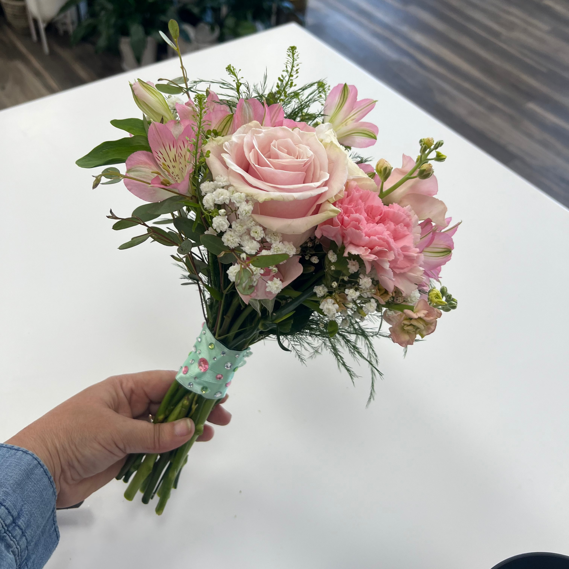 A hand holding a small bouquet of pink flowers, including a pink rose, pink carnations, and pink alstroemeria, with green foliage and white baby's breath, wrapped in a light green ribbon with a floral pattern. The bouquet is held over a white table in a flower shop with plants and a wooden floor in the background.
