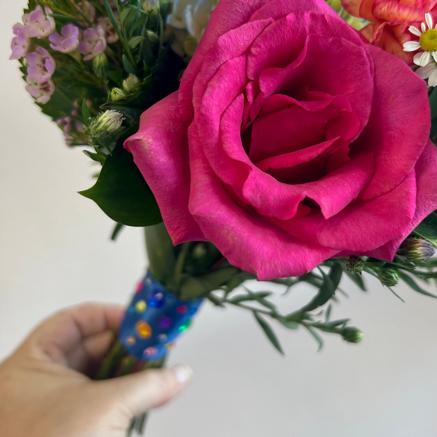 Flair Flower Bar in Florence, Kentucky features A close-up of a hand holding a vibrant flower Homecoming bouquet featuring a large magenta-pink rose, purple daisies, and other colorful flowers, with a blue glittery wrapped handle against a plain light background. 