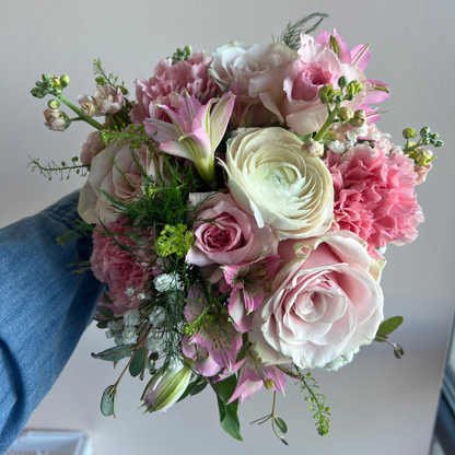A hand holding a vibrant homecoming floral bouquet featuring pink and white roses, carnations, lilies, and green foliage, with a person's blue denim sleeve visible on the left against a plain light gray wall. Perfect for Prom or Homecoming