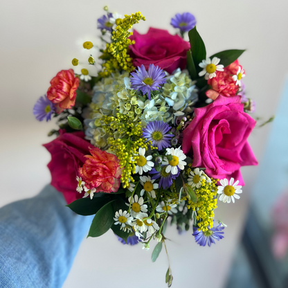 A vibrant mixed floral bouquet held by a hand wearing a light blue denim sleeve, featuring bright pink roses, coral carnations, purple daisies, white daisies, and green filler flowers against a soft, out-of-focus light background for Homecoming or Prom in Union, Burlington, Florence, Kentucky and Cincinnati High Schools