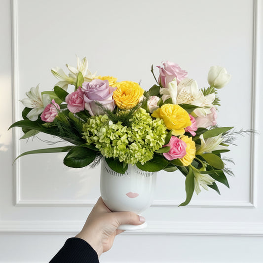 Arrangement of flowers with a pink ribbon held by a hand against a plain background