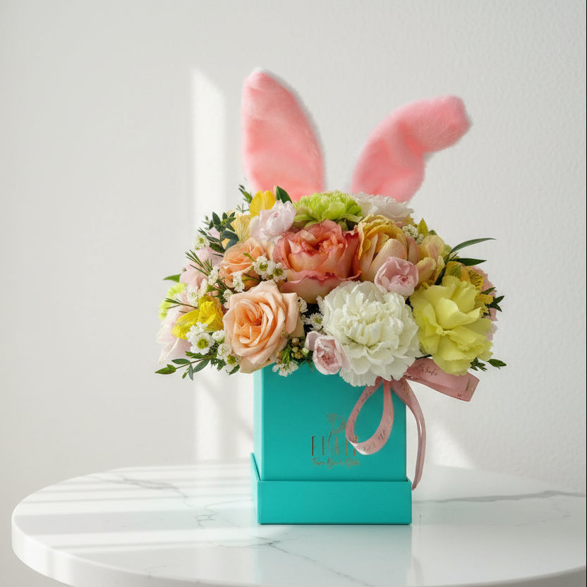 A floral arrangement featuring a variety of flowers with plush bunny ears on top, presented in a teal hat box with the brand name 'Flair'.