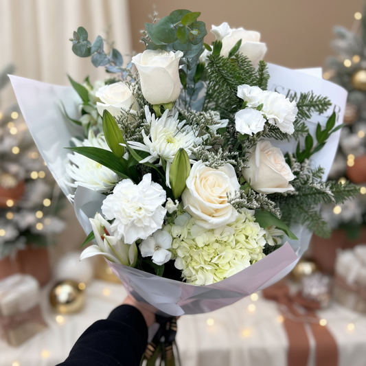 A bouquet of white roses, greenery, and accent flowers arranged to resemble a snow-kissed winter wonderland, held against a pink background.