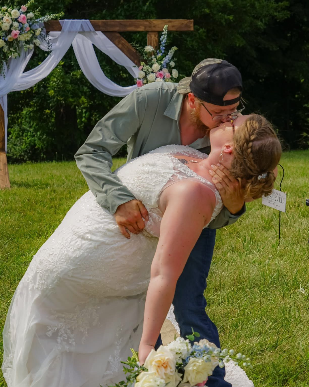 Man in a cap lifting a woman in a wedding dress at an outdoor wedding ceremony. Wedding Florist - Flair Flower Bar - Walton, Kentucky