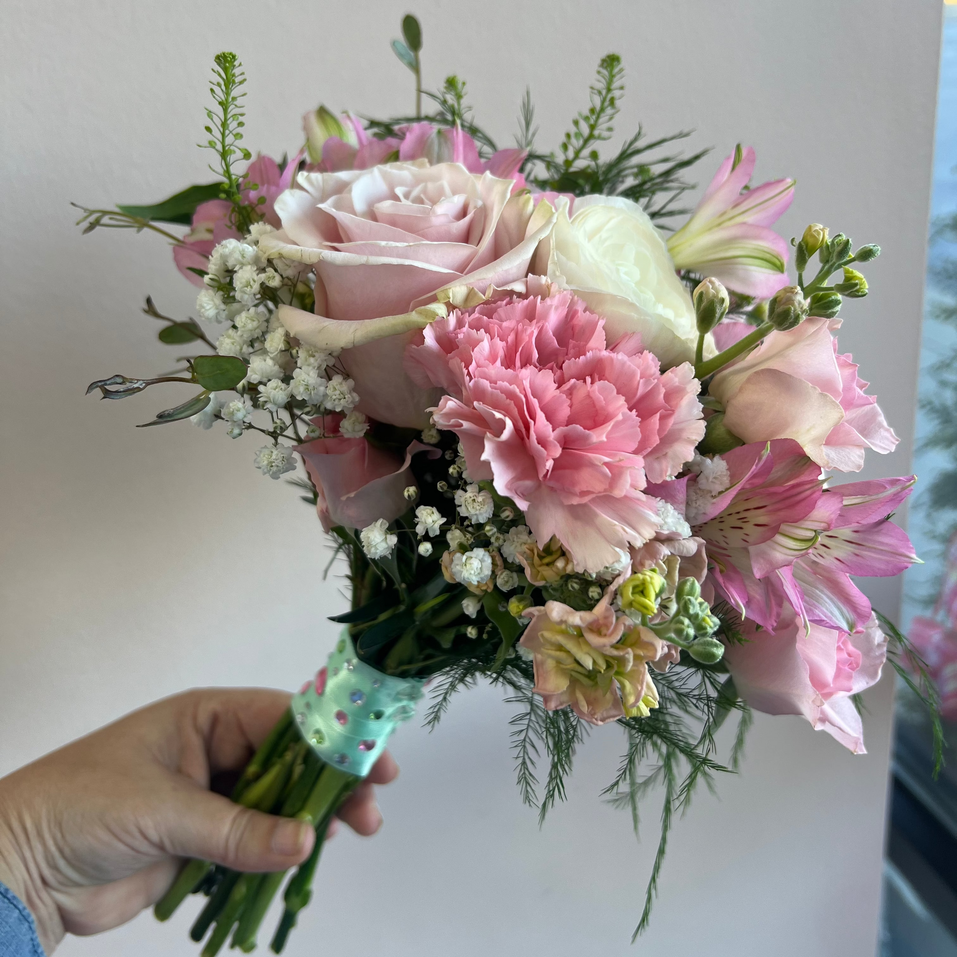 A hand holding a Prom bouquet of pink and white flowers, including roses, carnations, alstroemeria, and baby's breath, wrapped in a green and Rhinestone ribbon against a plain light gray wall in Florence, Kentucky designed by Flair Flower Bar